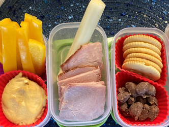 Tupperware storage containers filled with hummus, crackers, cheese, fruit, sliced ham and chocolate covered peanuts