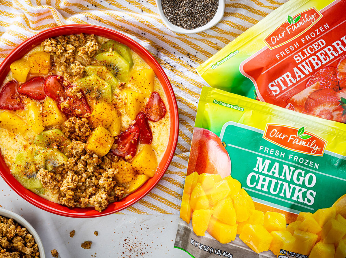 Photo of Our Family brand frozen mango chunks and frozen strawbers along side of a tropical smooth bowl with mangos, strawberries kiwi and granola