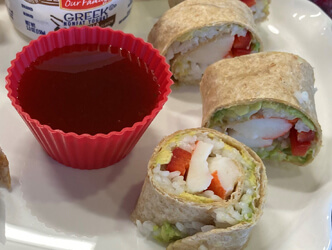 Photo of whole wheat tortilla wrap filled with avocado, imitation crab, rice, shredded carrots and red bell pepper alongside soy dipping sauce